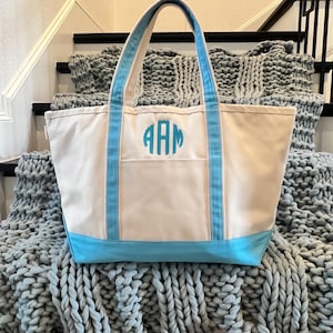 May include: A white canvas tote bag with blue trim and a monogram of the letters "AAM" in turquoise. The bag is sitting on a gray knitted blanket.