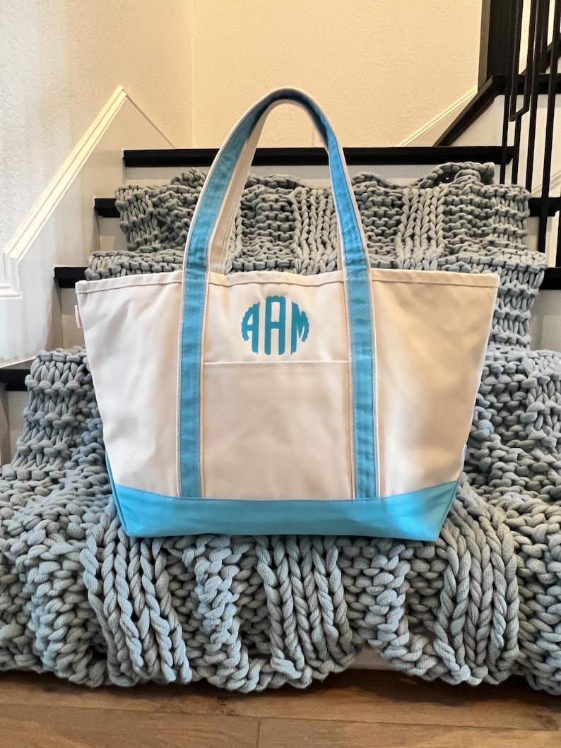 May include: A white canvas tote bag with blue trim and a monogram of the letters "AAM" in turquoise. The bag is sitting on a gray knitted blanket.