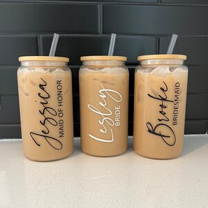 May include: Three clear glass tumblers with bamboo lids and straws. The tumblers are filled with a light brown liquid. Each tumbler has a personalised name and title: "Jessica Maid of Honour", "Lesley Bride", and "Brooke Bridesmaid".