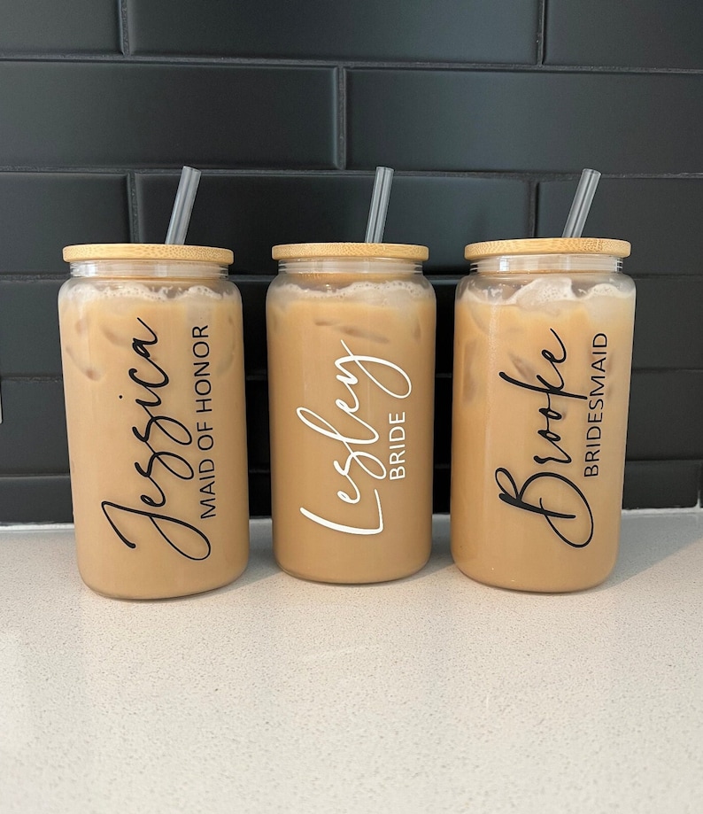 May include: Three clear glass tumblers with bamboo lids and straws. The tumblers are filled with a light brown liquid. Each tumbler has a personalised name and title: "Jessica Maid of Honour", "Lesley Bride", and "Brooke Bridesmaid".