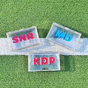 Pode incluir: Tr&ecirc;s caixas de acr&iacute;lico transparentes com letras vermelhas e azuis que soletram "SNR", "MD" e "KDP". As caixas est&atilde;o sobre uma superf&iacute;cie de grama artificial verde.