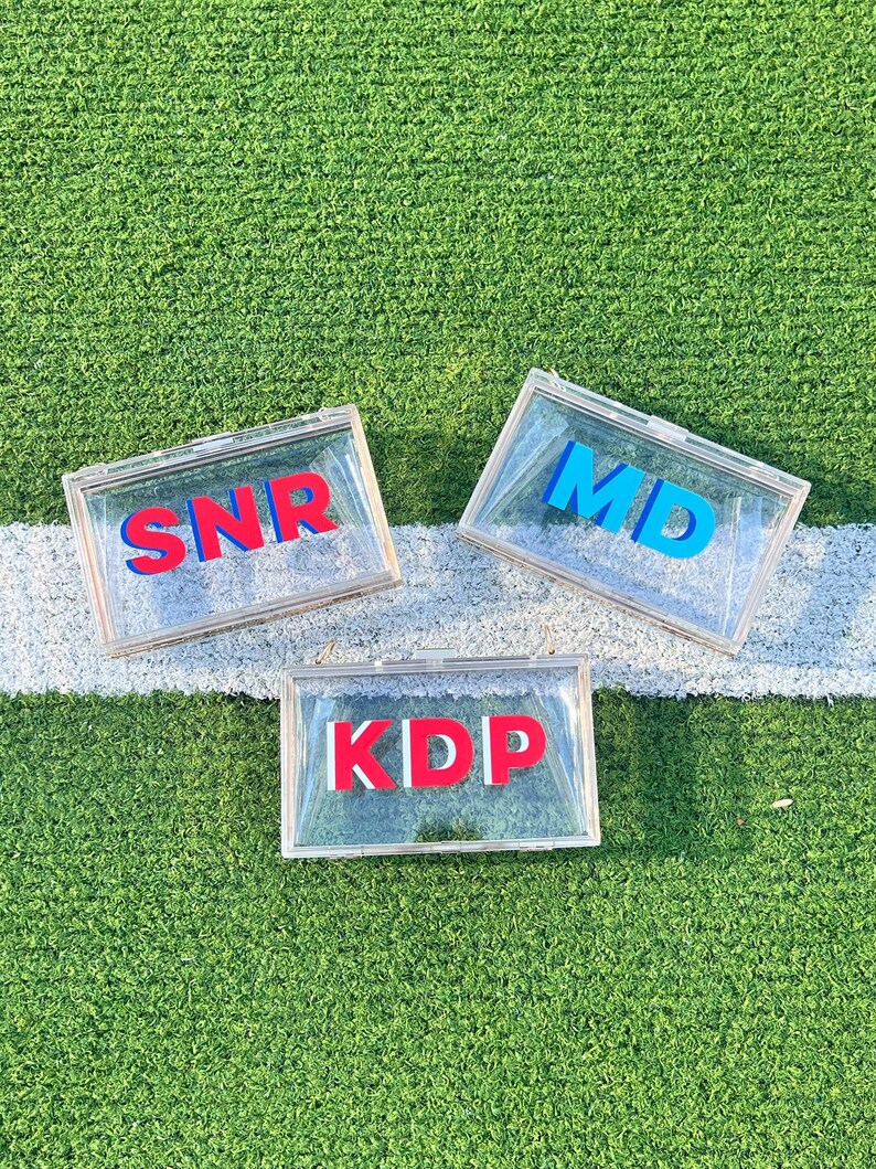 Pode incluir: Tr&ecirc;s caixas de acr&iacute;lico transparentes com letras vermelhas e azuis que soletram "SNR", "MD" e "KDP". As caixas est&atilde;o sobre uma superf&iacute;cie de grama artificial verde.