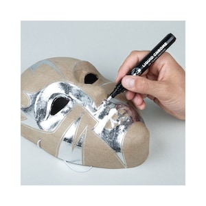 May include: A brown paper mache mask with silver metallic accents is being detailed with a black Liquid Chrome marker. The mask has eye and mouth openings and a white string for wearing. The marker is labeled "Liquid Chrome 20 Years Edition."