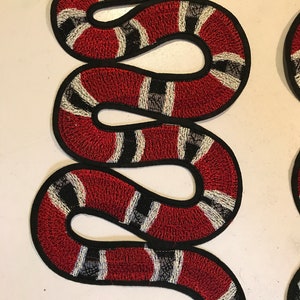 Può includere: Un toppa ricamata a forma di serpente rosso, bianco e nero. Il serpente è arrotolato a forma di 'S' con la testa in cima alla toppa.