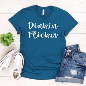 May include: Teal t-shirt featuring the white text "Dinkin Flicka". The image also shows denim shorts and white canvas trainers. A small green plant is also visible.
