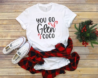 以下が含まれることがあります： 白いTシャツに、黒い文字で「You go Glen Coco」と書いてあり、赤と白のキャンディーケーンのグラフィックが描かれています。