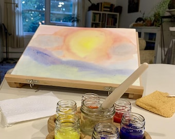 May include: A wooden paint organizer holding five small glass jars filled with yellow, blue, and red liquid paint. A paintbrush rests in one jar. Behind the organizer sits a wooden easel holding a watercolor painting of a yellow sun in a sky.
