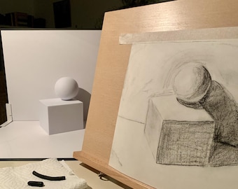 May include: A wooden easel holds a charcoal drawing of a sphere resting on a cube. Beside the easel, a white sphere sits on a white cube. Two pieces of black charcoal lie on a white paper towel in the foreground, completing the still life setup.