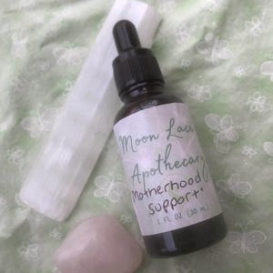 May include: A dark amber glass dropper bottle labeled Moon Lace Apothecary Motherhood Support 1 fl oz (30 ml), accompanied by a white selenite crystal wand and a smooth, pale pink rose quartz stone, all arranged together.