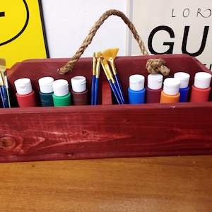 May include: A red wooden paint organizer with a rope handle. The organizer has eight slots for paint bottles and a space for paintbrushes. The organizer is filled with eight bottles of paint in various colors and four paintbrushes.