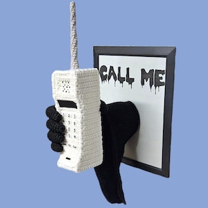 May include: A white crocheted cellular phone with a gray antenna is held by a black crocheted hand. The phone has a keypad and is positioned in front of a framed sign that reads "CALL ME" in black dripping letters.
