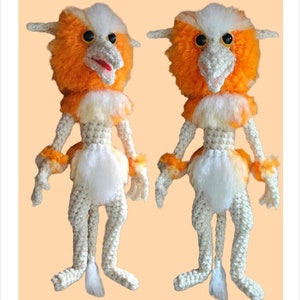 May include: A crocheted plush toy with orange fur, white fur, and black eyes. The toy is standing on a light orange background. The toy is called "Fire Dancer".