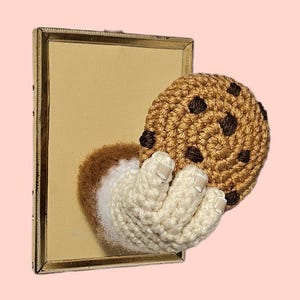 May include: A rectangular gold-toned mirror frame featuring a three-dimensional crochet element attached to the surface. The crochet piece depicts a cream-colored hand emerging from the mirror, holding a round, tan, chocolate chip cookie.
