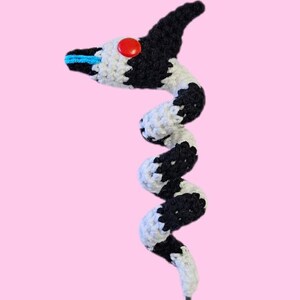 Op de afbeelding: Een handgemaakt gehaakt slangenknuffel in zwart en wit met een rode knoop als oog en blauwe accenten. De slang heeft een lange, grijze staart en staat tegen een roze achtergrond.