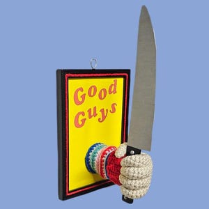 May include: A novelty art piece featuring a yellow sign with the words "Good Guys" in red, a crocheted hand, and a large knife. The sign is framed in black and red, with a light blue background.