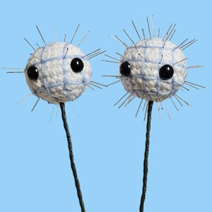 May include: Two whimsical, handmade, white crochet spheres with black eyes and metal pin accents. The spheres are connected to dark green stems, set against a light blue background. The crochet design features a grid pattern with light blue yarn.