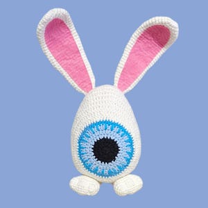 May include: A whimsical, handmade crochet bunny with a unique eye design. The white bunny features long ears with pink inner ears, a large blue and black eye, and small feet. The plush toy is set against a light blue background.