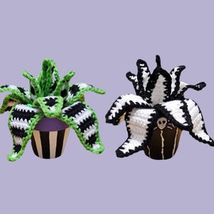 May include: Two crochet plant decorations. One has green and black striped leaves with a purple and black striped pot. The other has black and white striped leaves with a brown pot and a skull design.