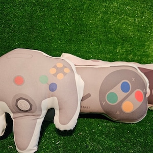 May include: Three plush pillows shaped like classic video game controllers. The pillows are gray, white, and black with colorful buttons. The first pillow is a gray Nintendo 64 controller, the second is a black Super Nintendo controller, and the third is a gray Nintendo GameCube controller.