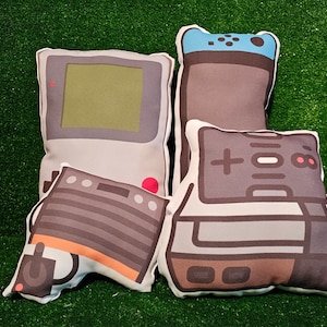 May include: Four plush pillows shaped like retro video game consoles. The pillows are gray, brown, and white with black details. The pillows feature a Game Boy, a Nintendo Entertainment System, a Super Nintendo Entertainment System, and a Sega Genesis.
