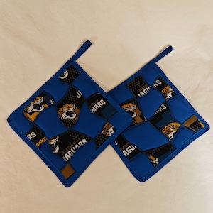 May include: Two blue and black quilted pot holders with the Jacksonville Jaguars logo. The pot holders are square with a loop for hanging. The logo features a jaguar head and the team name.