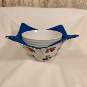 May include: A white ceramic bowl sits inside a blue and white fabric bowl cozy. The cozy has a star shape and is decorated with a repeating pattern of red, white, and blue basketballs.