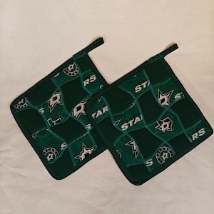 May include: Two square fabric pot holders in dark green featuring a repeating pattern of the Dallas Stars hockey team logo and the text "STARS" and "DALLAS". Each pot holder has a small fabric loop at the corner for hanging in the kitchen.
