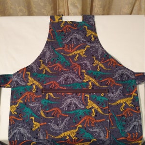 May include: A gray apron with a dinosaur skeleton print in yellow, orange, green, and blue. The apron has a brown leather pocket.