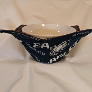 May include: A black and green fabric bowl cozy with the Philadelphia Eagles logo and the words "Eagles" and "AFL" printed on it. The cozy is holding a white ceramic bowl.