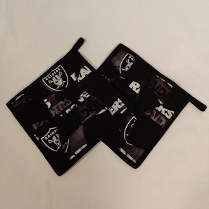 May include: Two black and white pot holders with the Las Vegas Raiders logo and the word "Raiders" repeated on them.
