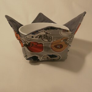 May include: A gray fabric bowl cozy with a football helmet print. The cozy is folded and a white bowl is inside.