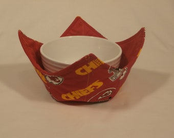 May include: Red fabric bowl cozy with a white bowl inside. The fabric has a pattern of yellow "Chiefs" text and a football player graphic.