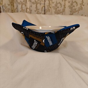 May include: A bowl cozy featuring a Jacksonville Jaguars team logo design. The fabric is black, blue, and gold, with the word "Jaguars" printed on it. The cozy is designed to hold a bowl and keep it warm.