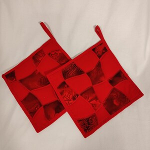 May include: Two red quilted pot holders with a patchwork design. The pot holders have a loop handle on each side.