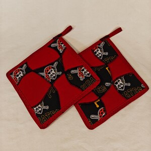 May include: Two square fabric pot holders featuring a patchwork design of solid red fabric and black fabric printed with a pirate logo and the text Pirates. Each pot holder has a small fabric loop at the corner for hanging and red stitched edges.
