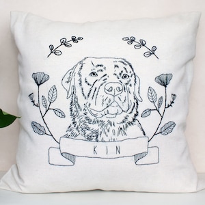 May include: A white pillow with a black embroidered image of a dog's head with floral accents. The dog's name 'KIN' is embroidered below the image.