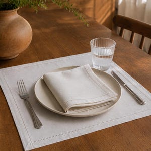 May include: A square light beige placemat with a stitched border, featuring a matching folded napkin on a round ceramic plate. A stainless steel fork and knife are placed on the sides, accompanied by a clear ribbed glass of water.