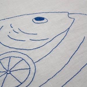 May include: A white fabric featuring blue thread embroidery of a fish and a citrus slice. The fish design includes a solid blue circular eye and curved lines for gills, while the citrus slice is depicted with a central point and radial lines.