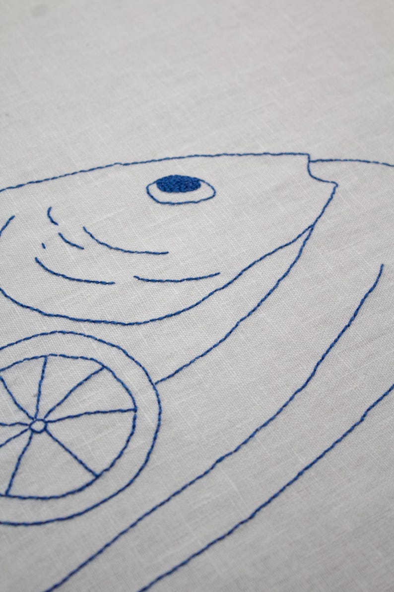 May include: A white fabric featuring blue thread embroidery of a fish and a citrus slice. The fish design includes a solid blue circular eye and curved lines for gills, while the citrus slice is depicted with a central point and radial lines.