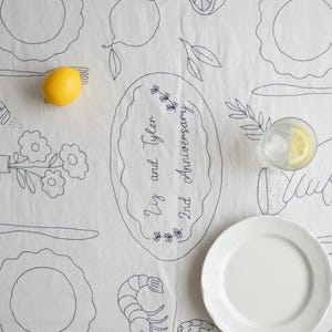 May include: A white fabric tablecloth featuring black line art illustrations of place settings, flowers, a shrimp, an artichoke, and lemons. The center includes an oval frame with the text Liz and Tyler 2nd Anniversary. A yellow lemon sits on the cloth.
