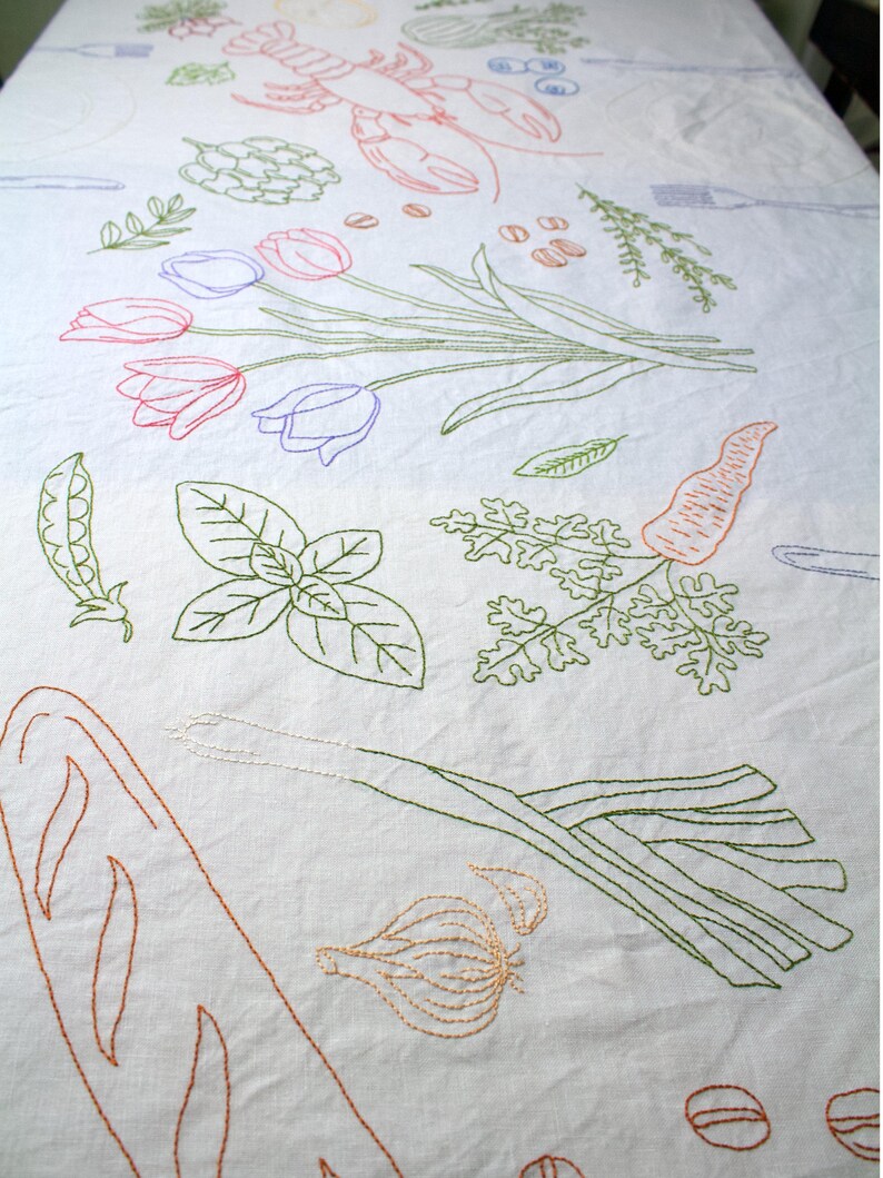 May include: A white tablecloth featuring colorful embroidered line art illustrations of food items including a lobster, tulips, a carrot, a leek, garlic, herbs, peas, and bread, interspersed with outlines of forks and knives across the fabric surface.
