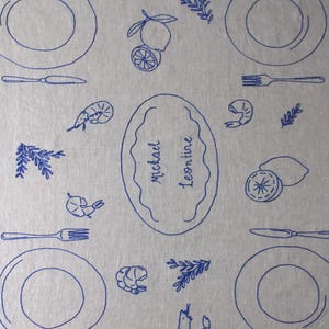 May include: A light gray fabric placemat featuring a repeating blue line-art pattern of plates, forks, knives, shrimp, lemons, and sprigs of herbs. A central oval frame contains the handwritten text "Michael Leontine" in blue ink.