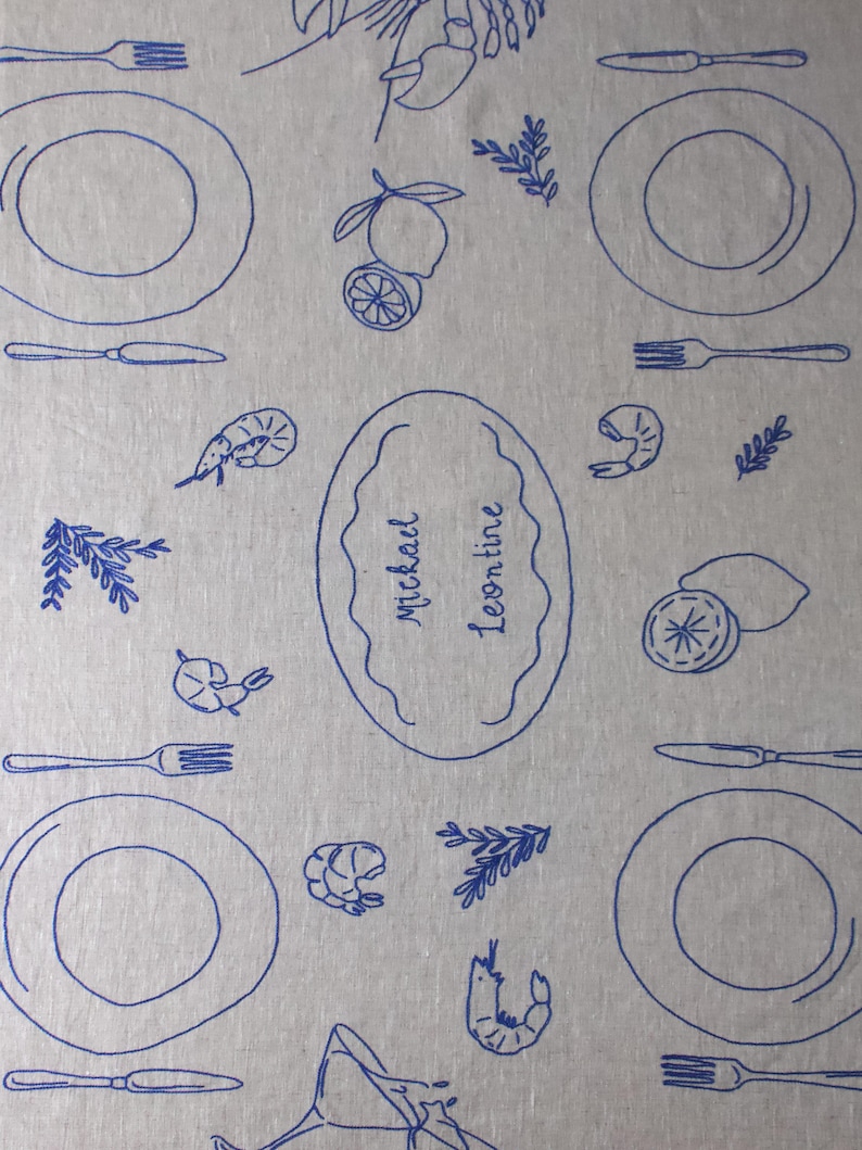 May include: A light gray fabric placemat featuring a repeating blue line-art pattern of plates, forks, knives, shrimp, lemons, and sprigs of herbs. A central oval frame contains the handwritten text "Michael Leontine" in blue ink.