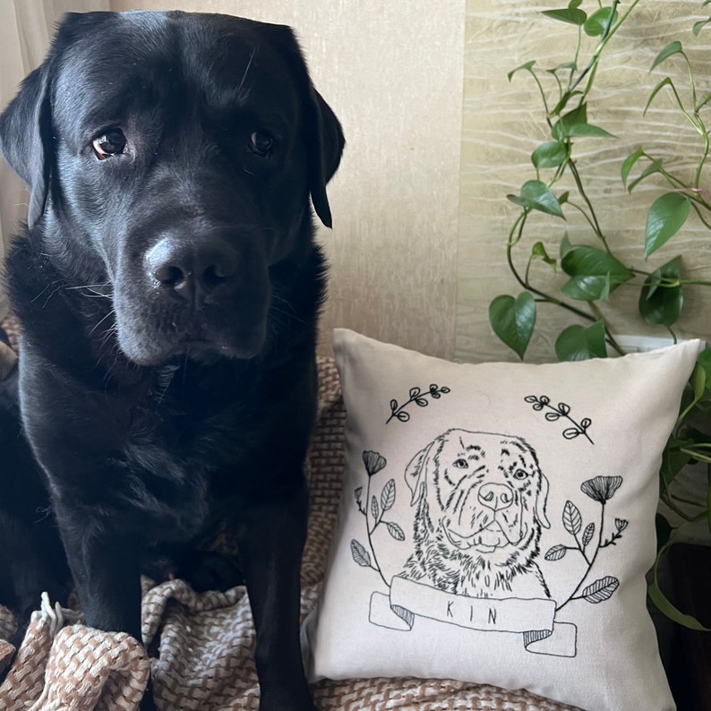 May include: A black Labrador Retriever sits next to a square, off-white pillow. The pillow features a black and white line drawing of a dog's face, surrounded by floral and leaf designs. The word "KIN" is written on a banner below the dog.
