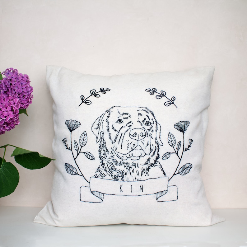 May include: A white pillow with a black embroidered image of a dog's head with floral accents. The dog's name 'KIN' is embroidered below the image.