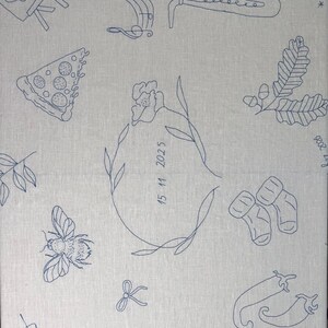 May include: A light gray fabric featuring dark blue line art embroidery of various icons including a saxophone, a pizza slice, a bee, baby socks, chili peppers, a martini glass, and a central floral wreath enclosing the date 15 11 2025.