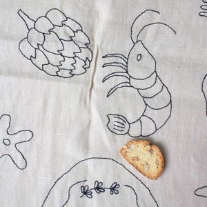 May include: A beige linen fabric featuring black embroidered line art of an artichoke, a shrimp, and a starfish. A single almond biscotti rests on the fabric near a partially visible embroidered oval frame containing the text "Liz 2nd".