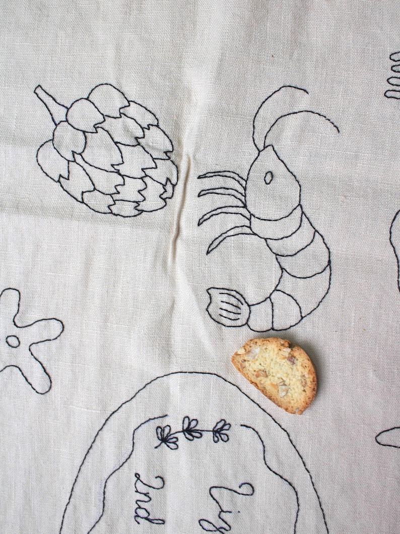 May include: A beige linen fabric featuring black embroidered line art of an artichoke, a shrimp, and a starfish. A single almond biscotti rests on the fabric near a partially visible embroidered oval frame containing the text "Liz 2nd".