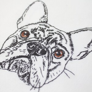 May include: Embroidered close-up of a French Bulldog's face. The dog's features are detailed with black thread on a white background. The dog has brown eyes and a black nose, with visible stitching creating the fur and facial details.