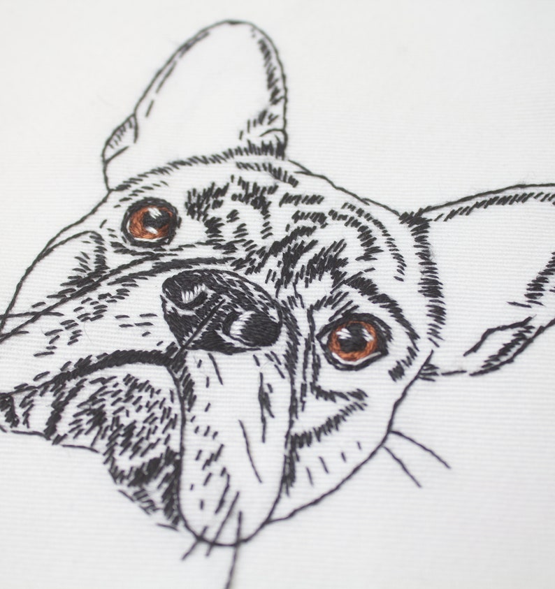 May include: Embroidered close-up of a French Bulldog's face. The dog's features are detailed with black thread on a white background. The dog has brown eyes and a black nose, with visible stitching creating the fur and facial details.